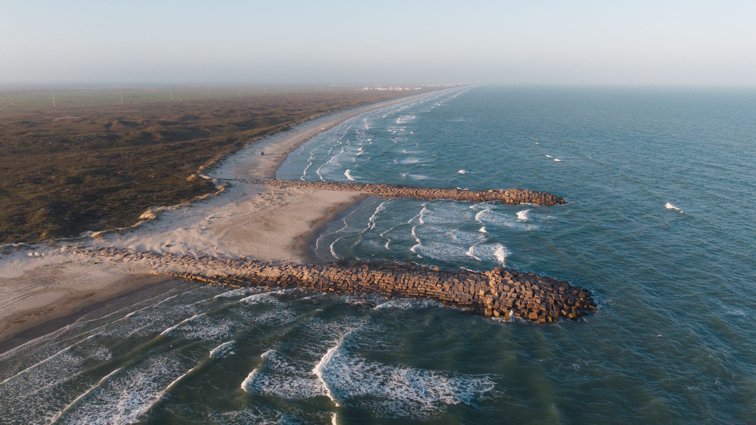 Aerial view of a Gulf beach with two rock jetties reaching into the surf and dunes behind the sand