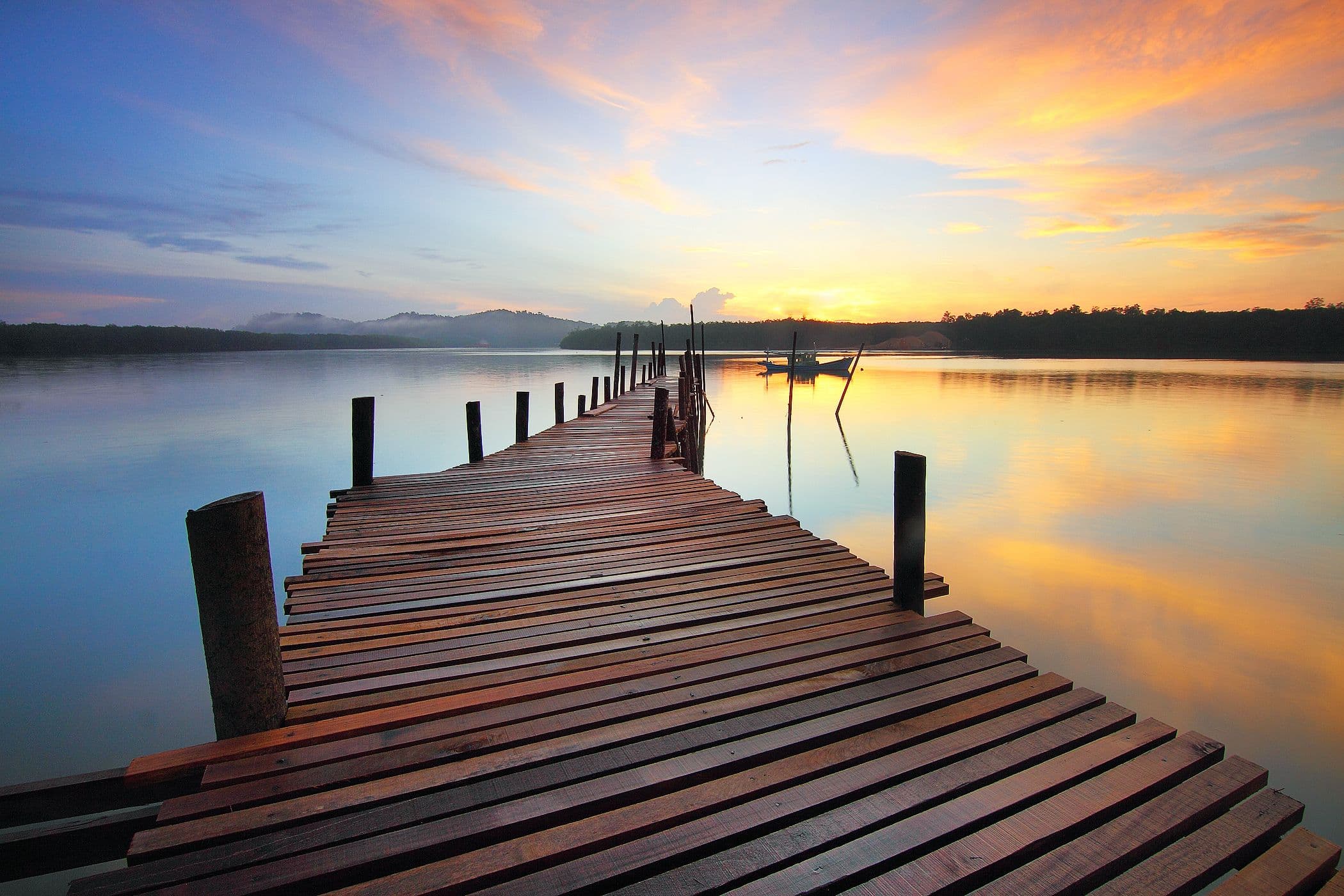 Wooden fishing pier over calm bay water framed by coastal trees at sunset